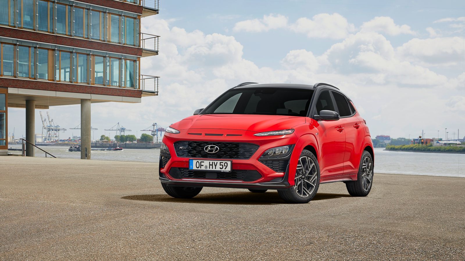 A red hyundai kona is parked in front of a building next to a body of water.