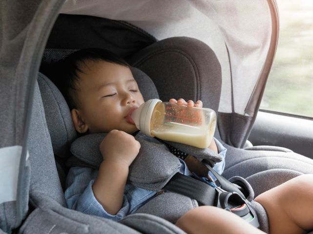 A baby is drinking milk from a bottle while sitting in a car seat. Kona Hibrid