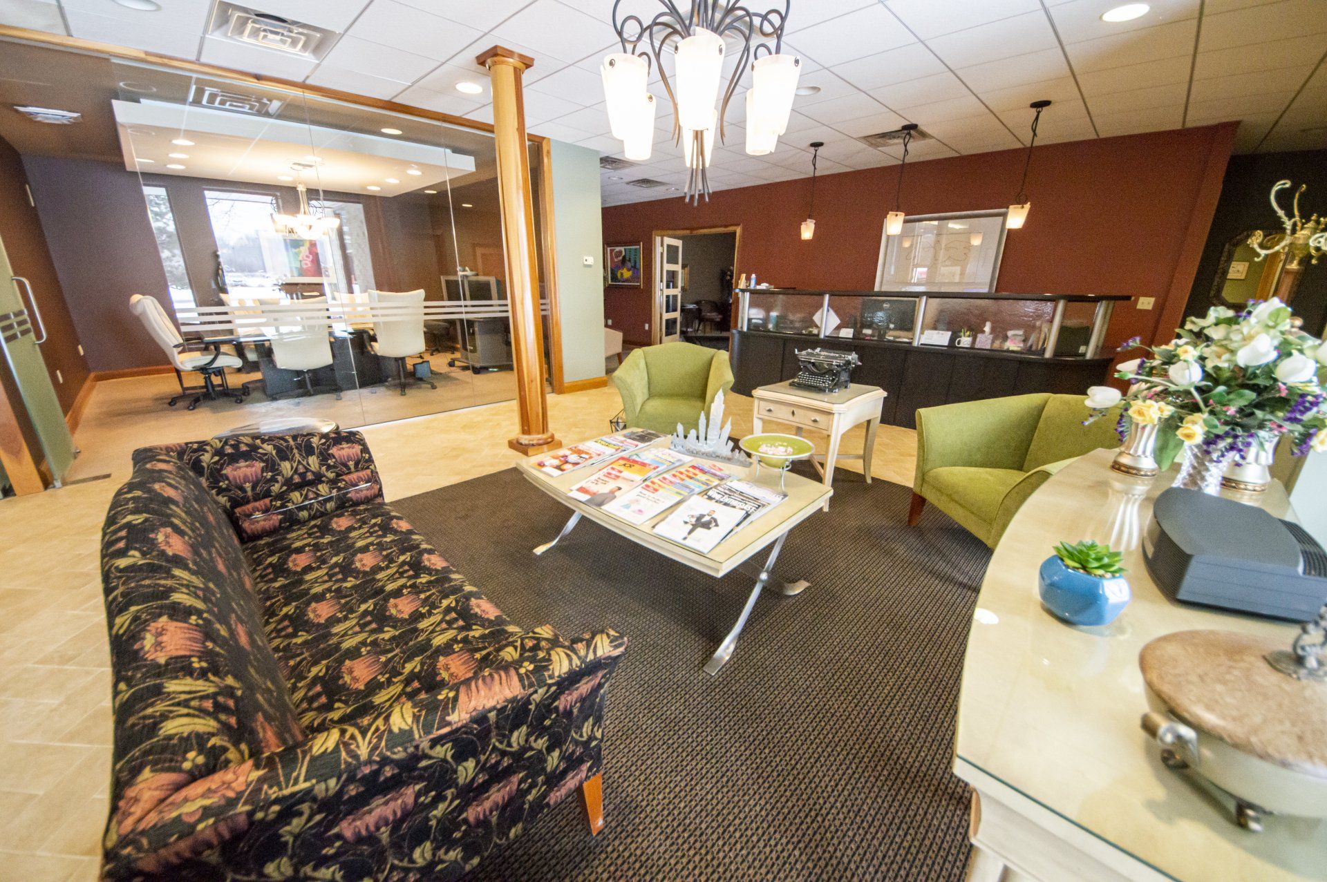 Lounge area in an office with floral sofa, green chairs, and a small table. Conference room in the background.