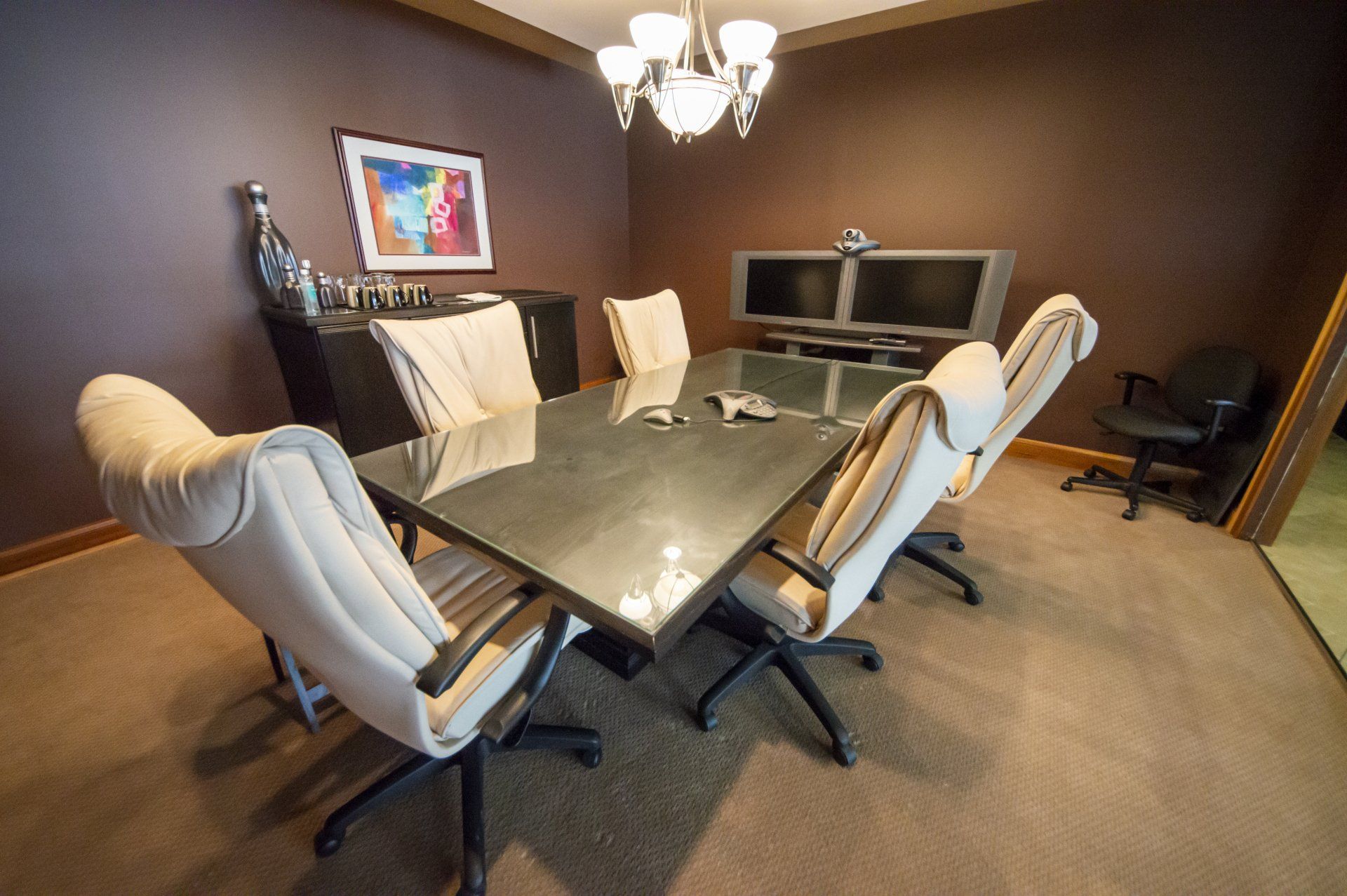 Conference room with a rectangular table and six cream-colored chairs, dark walls, and two monitors.
