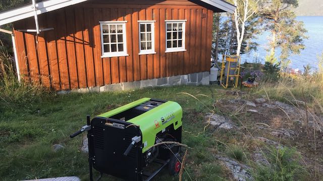 En bærbar, lysegrønn generator står i en gresskledd skråning foran en rustikk brun hytte nær et vann.