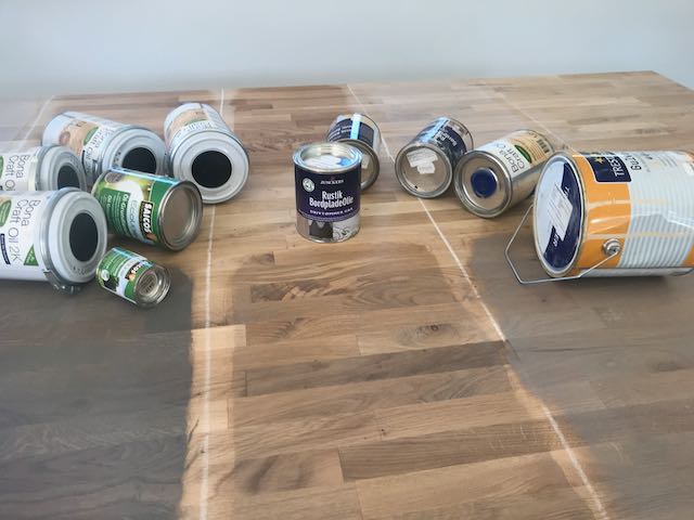 En samling av forskjellige malings- og trebehandlingsbokser spredt på et trebord med delvis beiset overflate.