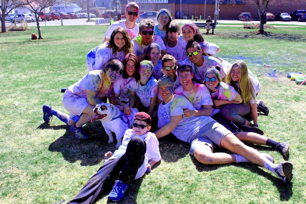 Group of people covered in colorful powder on a grassy field; some are smiling, some wearing sunglasses, and one has a dog.