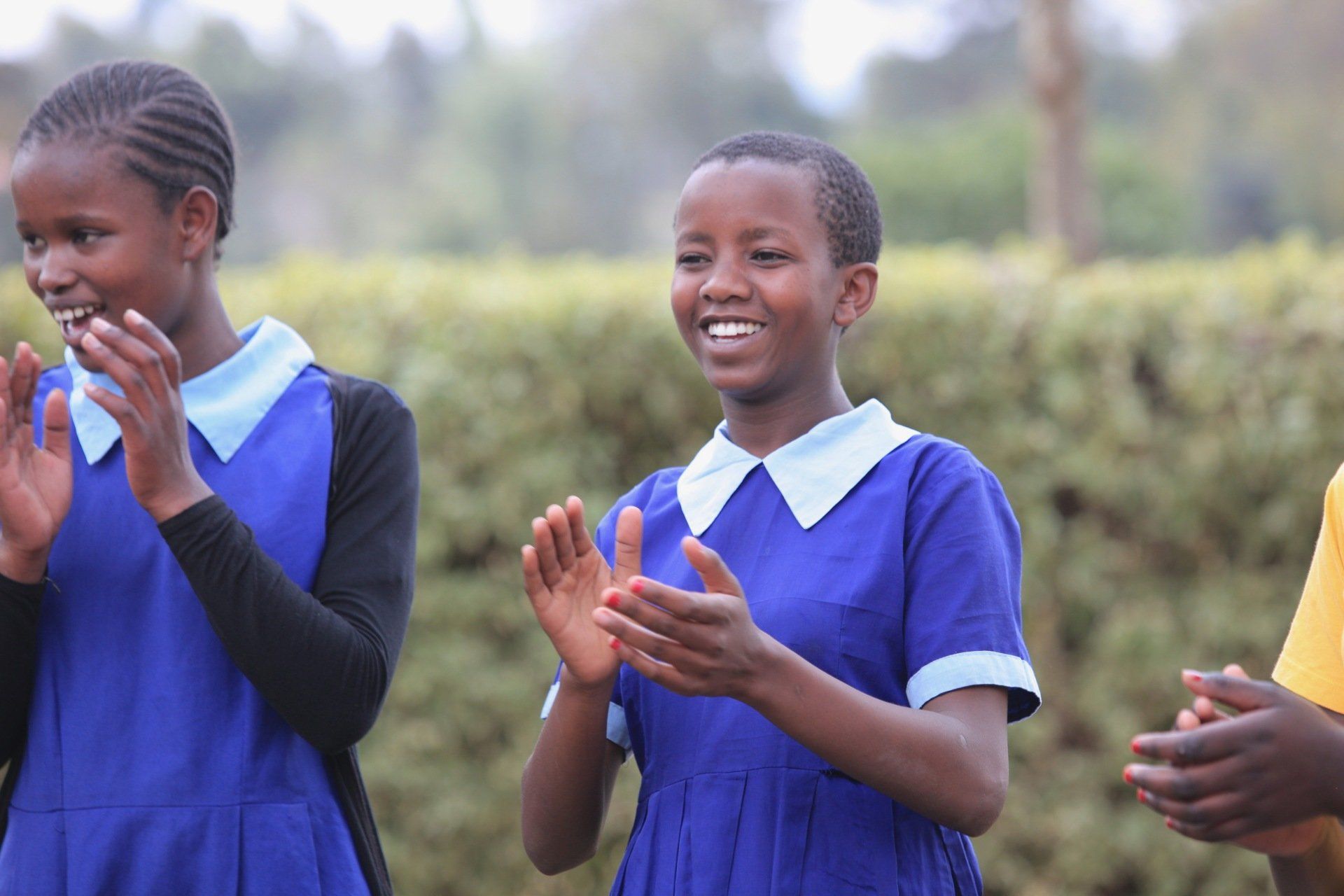 Two girls in blue school uniforms clapping, smiling outdoors.