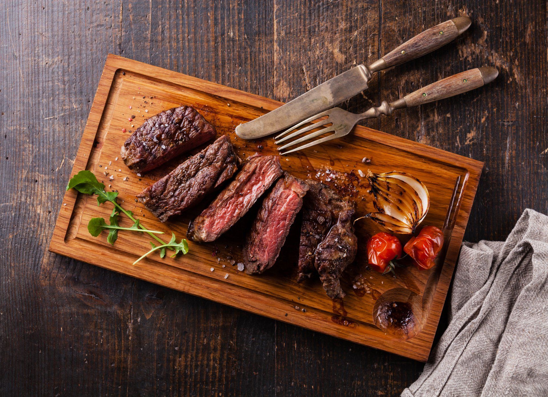 Sliced steak on wooden board with fork and knife, grilled vegetables.