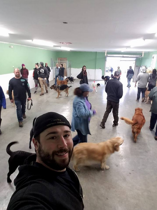 Man takes selfie in dog park with dogs and people, indoor setting.