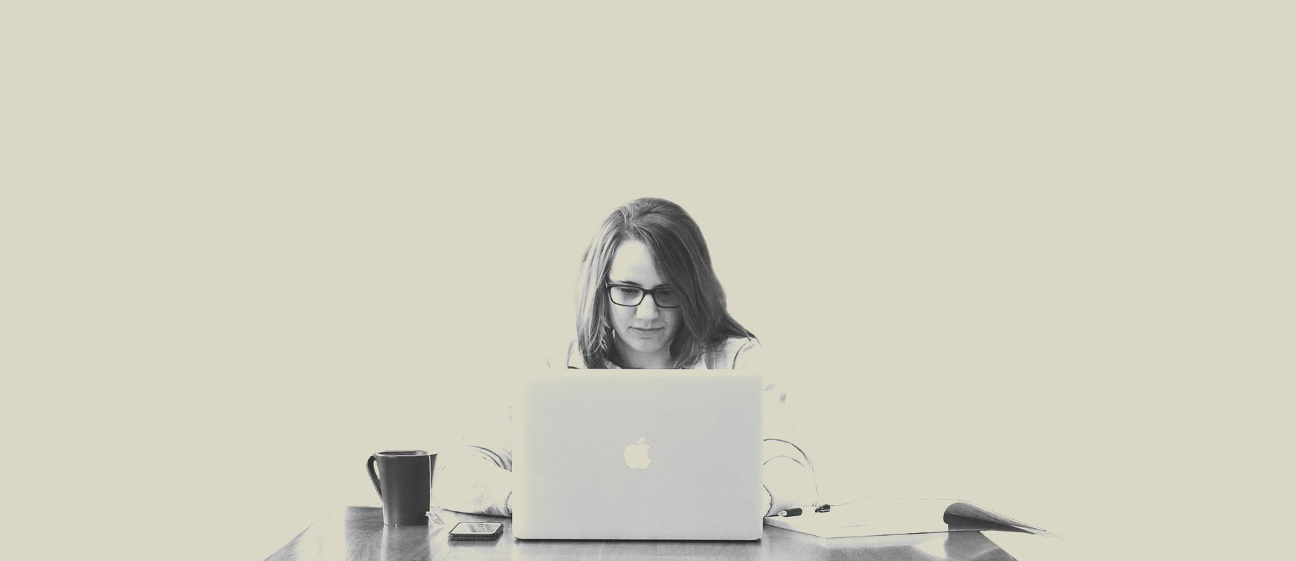 Woman working on a laptop at a desk with a coffee mug in a neutral-colored setting.