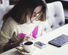 Eine Frau sitzt mit einer Tasse Kaffee und einem Handy an einem Tisch.