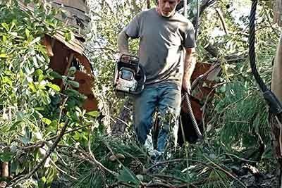 Man in jeans holds chainsaw, clearing debris near rusted metal structure, outdoors.