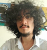 Homem de cabelos escuros cacheados e óculos, sorrindo levemente. Vestindo uma camisa clara, em ambiente interno.