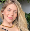 Mulher loira sorrindo, usando uma gargantilha dourada, com vegetação ao fundo.