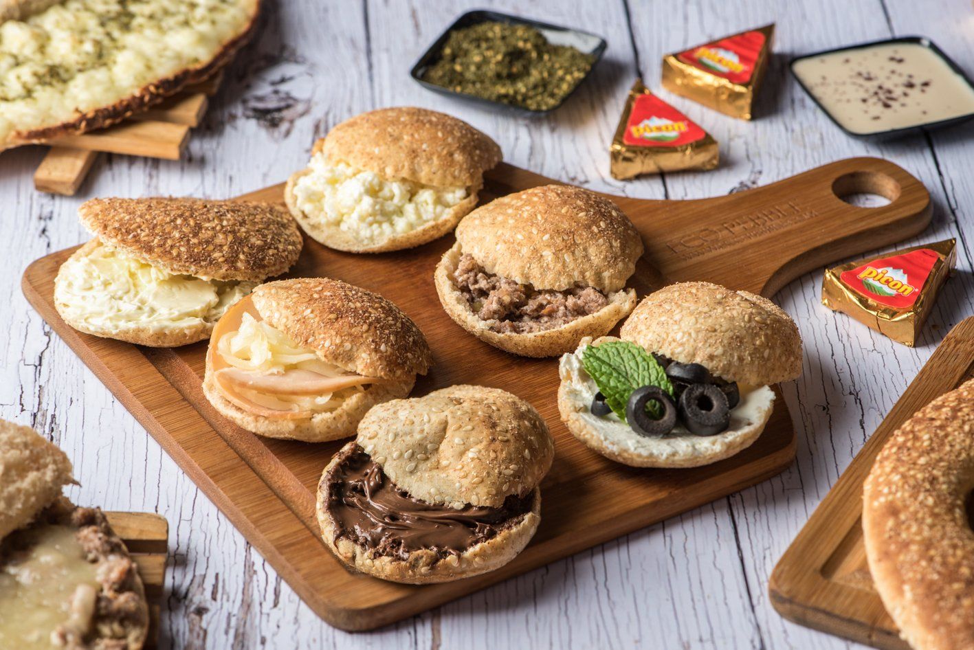 Small sandwiches on a wooden board, with various fillings like meat, olives, and chocolate.