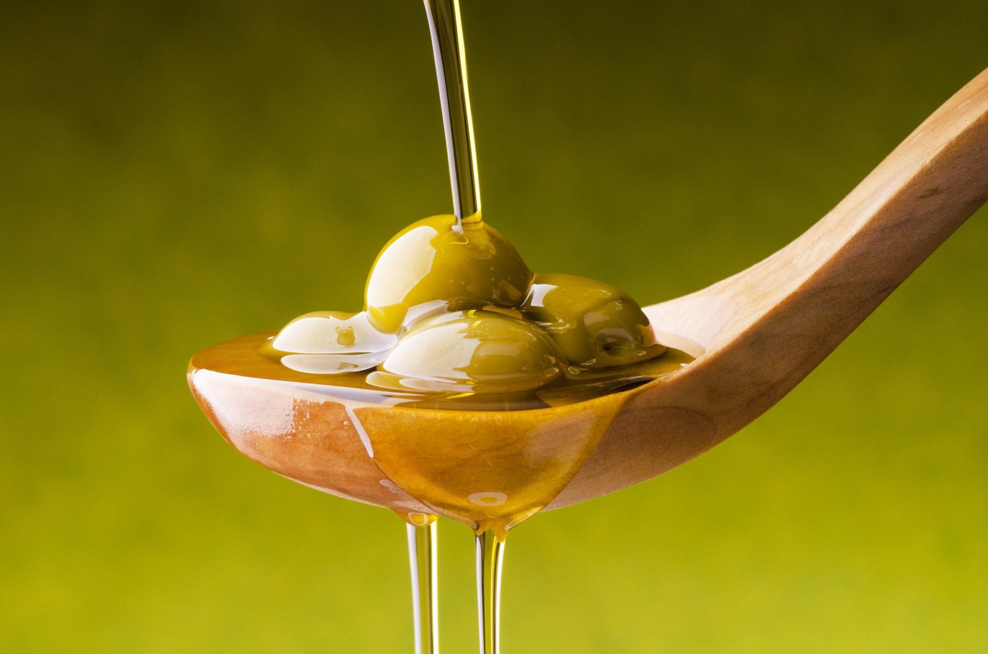 Green olives in a wooden spoon, olive oil pouring over them, against a yellow-green background.
