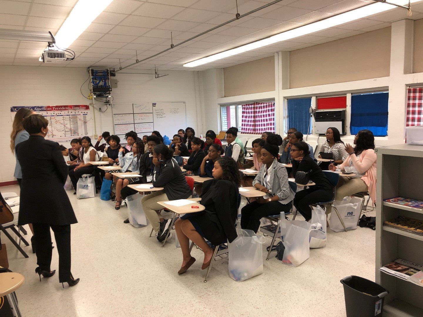 A woman is standing in front of a group of people sitting at desks in a classroom.