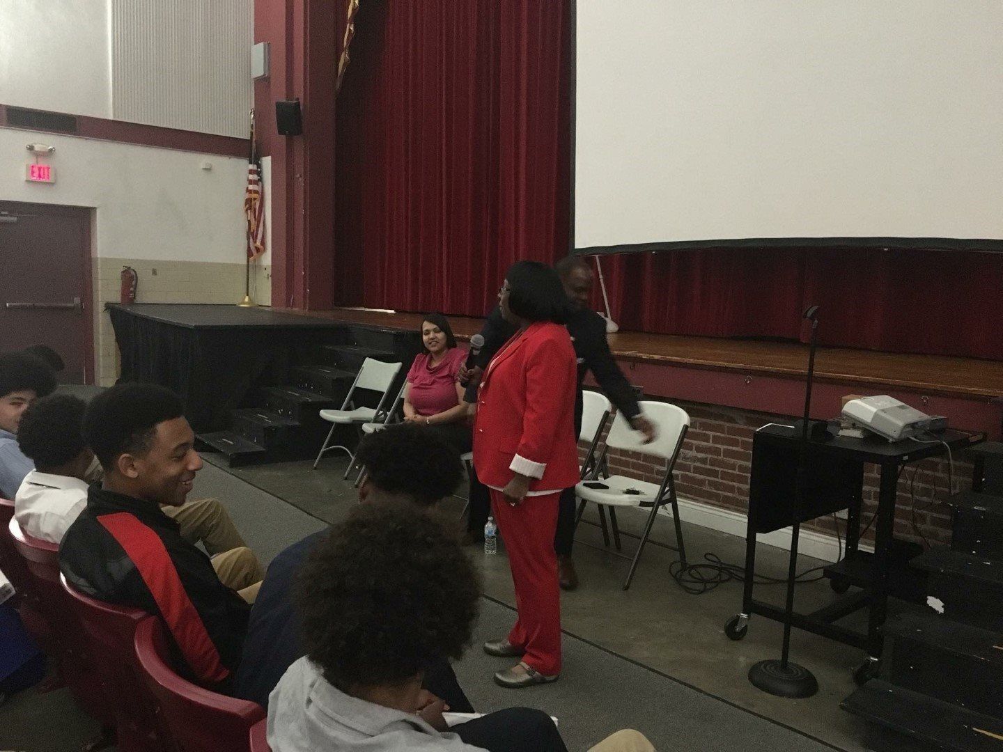 A woman in a red suit is standing in front of a group of people sitting in chairs.