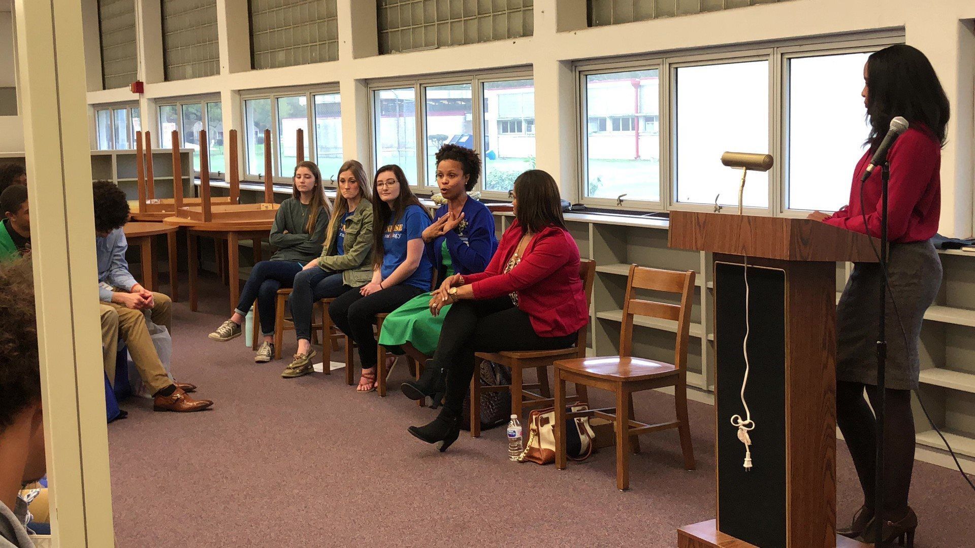 A woman is standing at a podium talking to a group of people sitting in chairs.