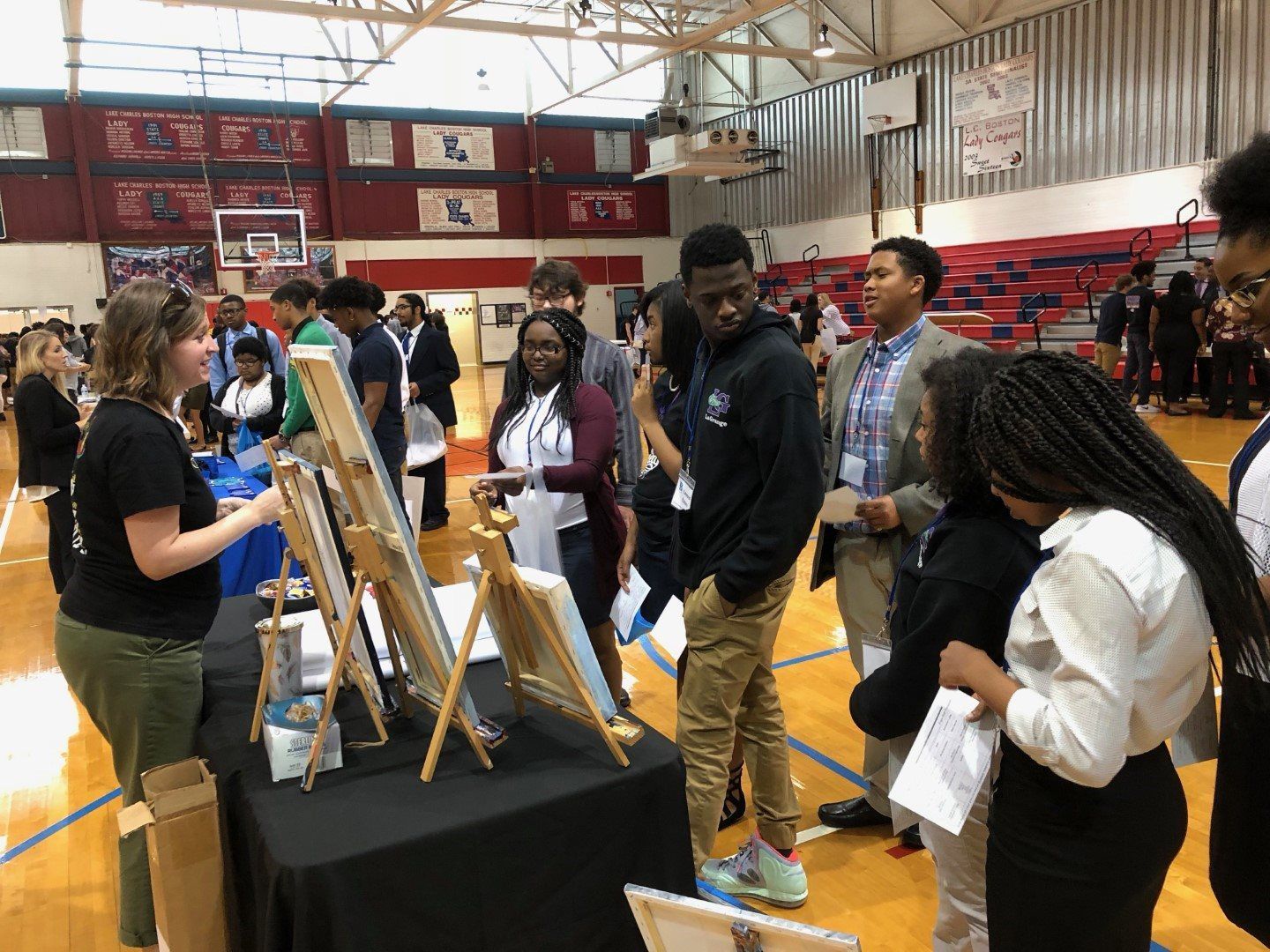A group of people are standing around a table in a gym looking at paintings on easels.