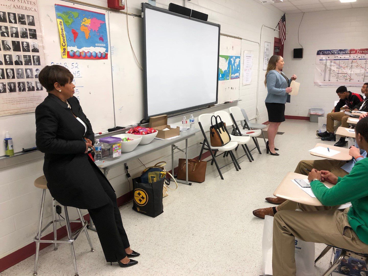 A woman is sitting on a stool in a classroom while a woman stands in front of a whiteboard.