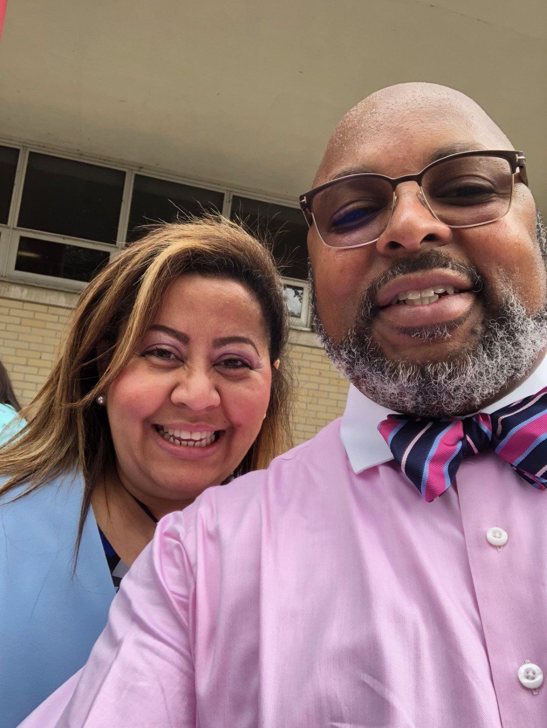A man and a woman are posing for a picture together . the man is wearing a pink shirt and a bow tie.