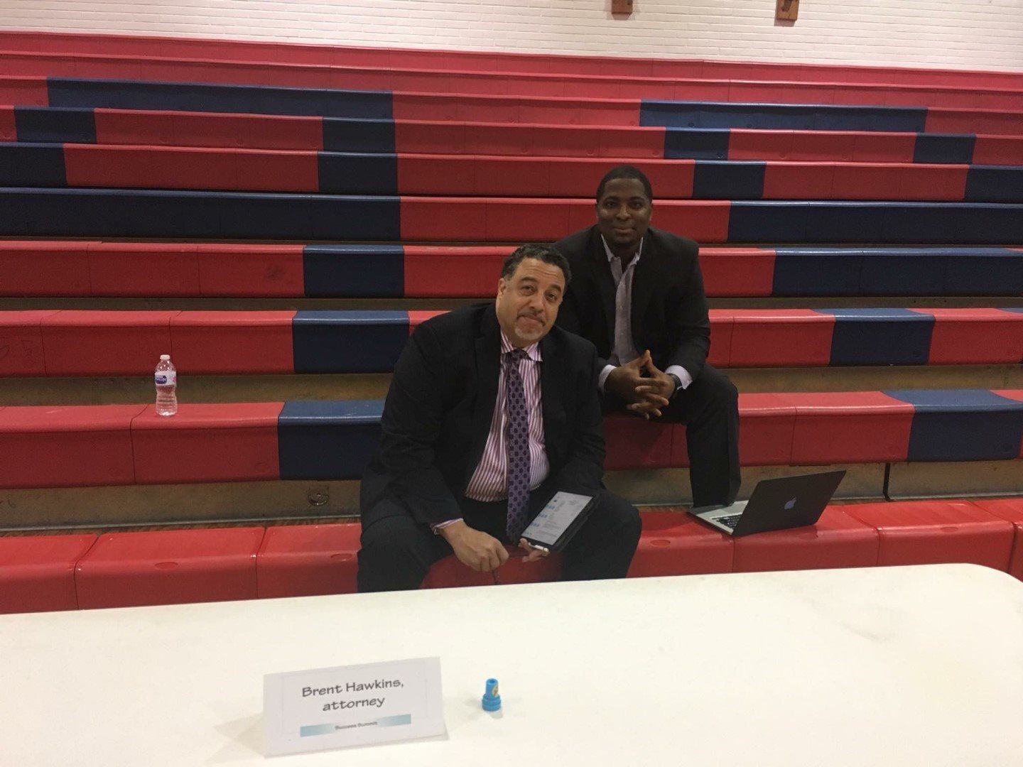 Two men are sitting on the bleachers in a gym with a laptop.