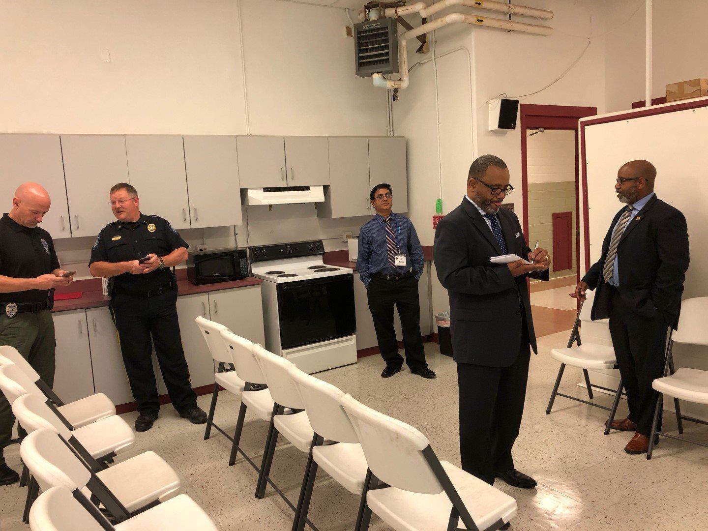A group of men are standing in a room with chairs and a stove.