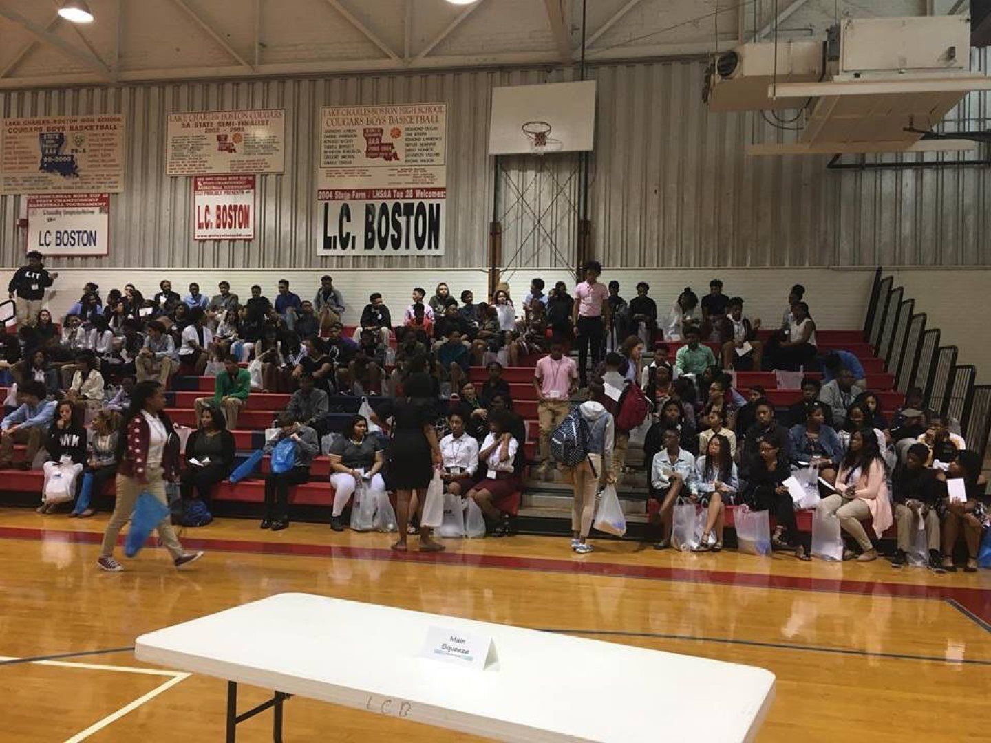 A large group of people are sitting in a gym with a sign that says l.c. boston