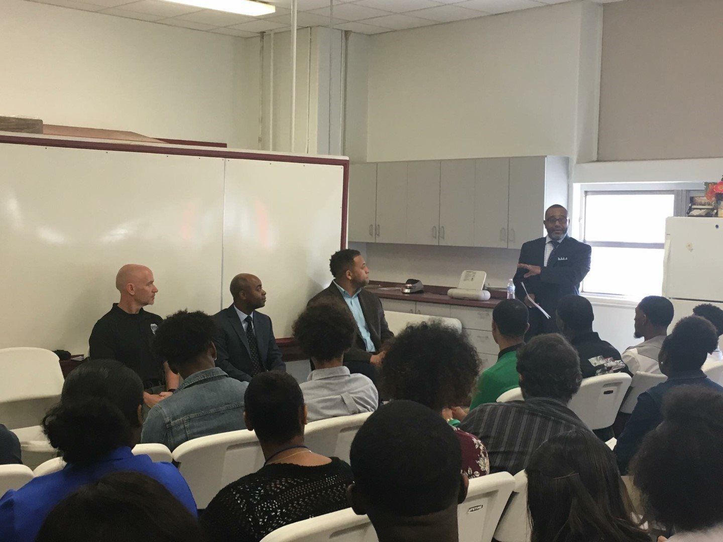 A group of people are sitting in a classroom with a man standing in front of them.