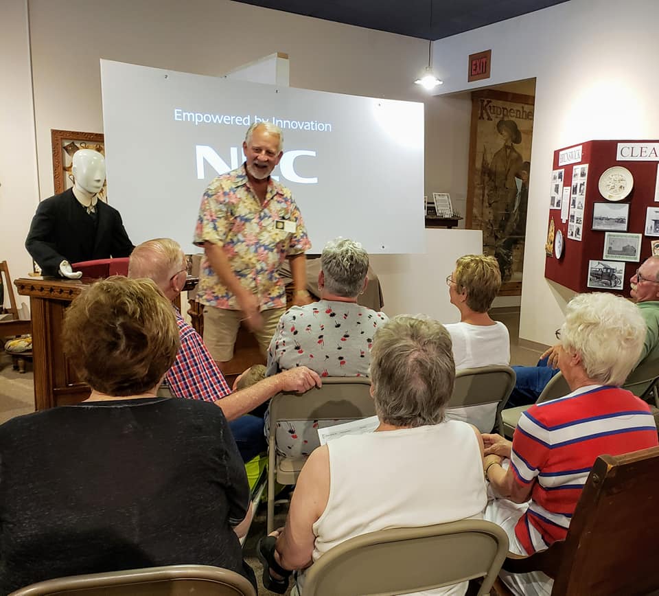Man speaking to seated audience in a room; presentation on screen, mannequin in suit.