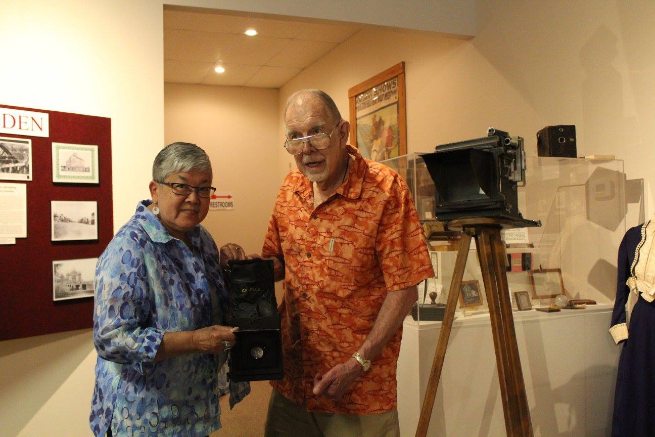 Two people holding a vintage camera in a museum, near a large camera on a tripod.