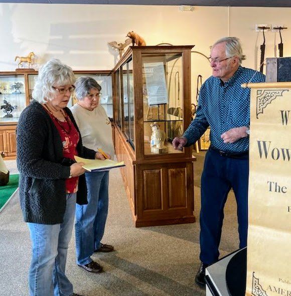 Three people in a museum look at a display case; one points and holds a sign.