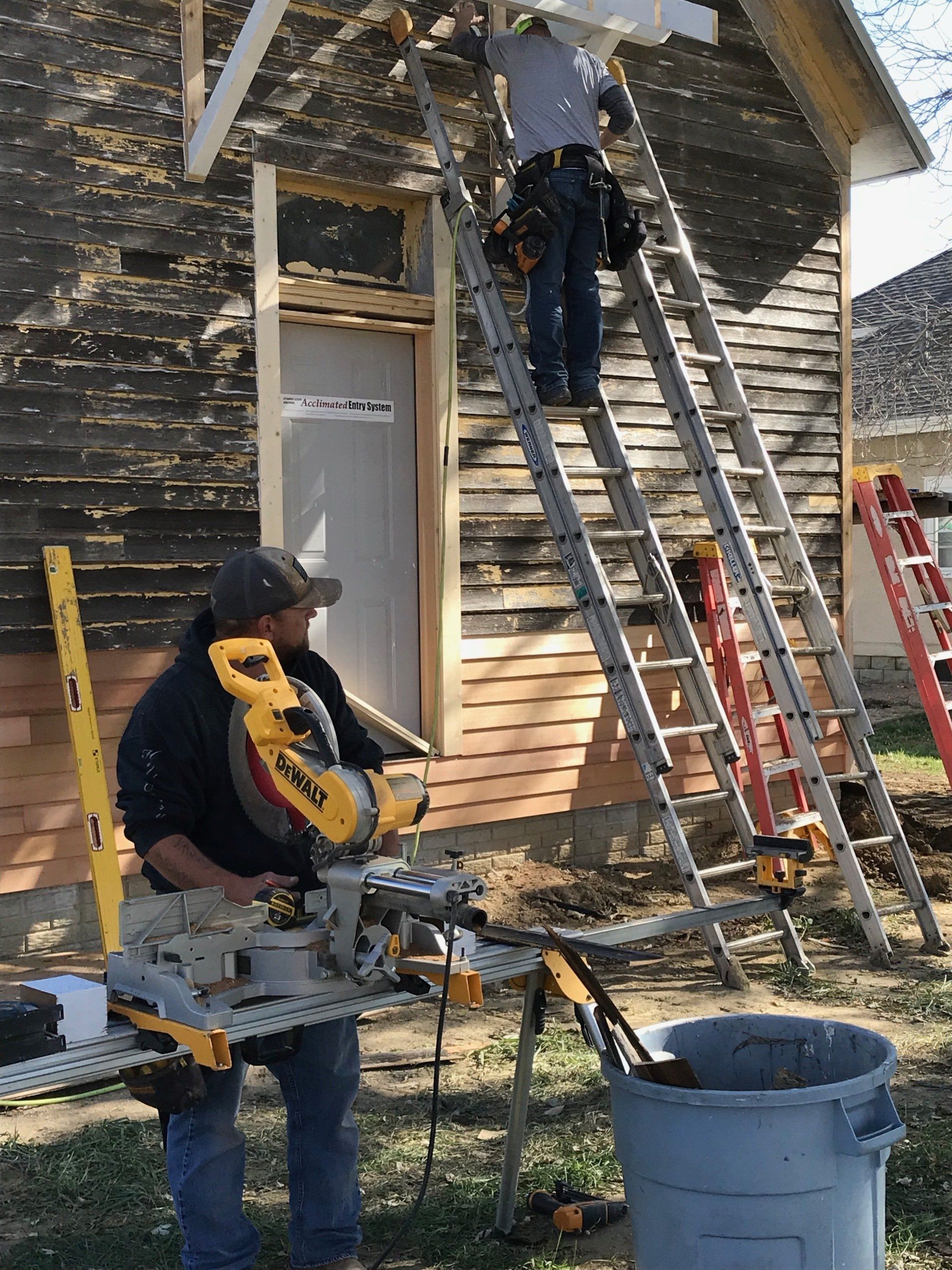 Two workers constructing a building exterior. One uses a saw, the other works on a ladder. Sunny, outdoor setting.