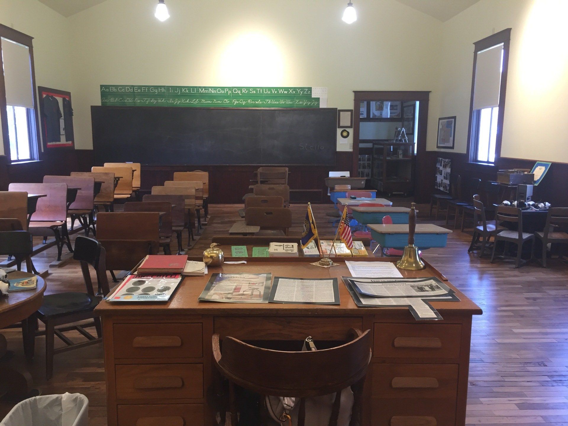Old school classroom, desks face blackboard, teacher's desk in foreground, windows on each side.