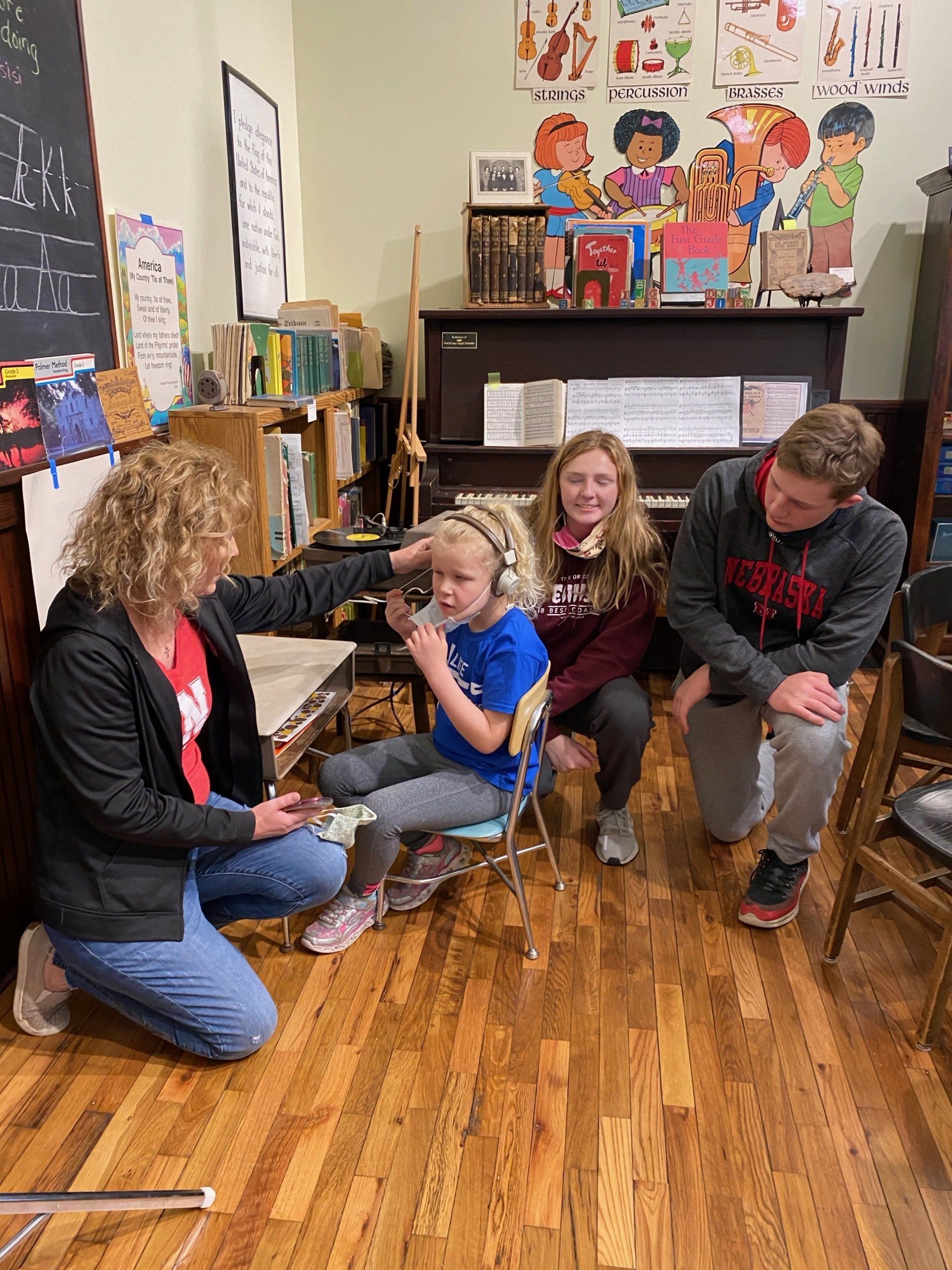 A woman adjusts headphones on a child seated on a stool, as two others watch near a piano in a room with books.
