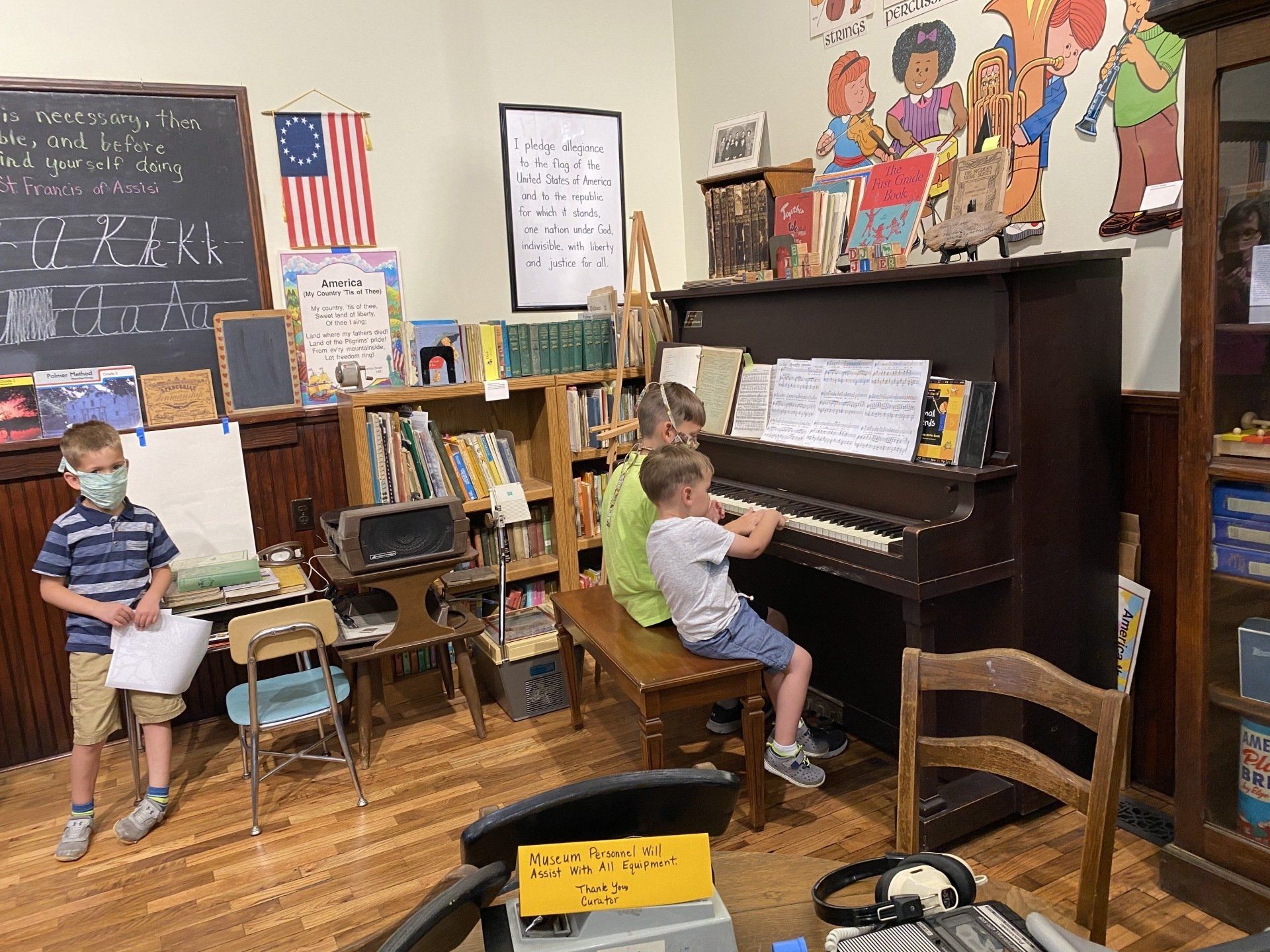 Children in a vintage classroom: One plays piano, others stand nearby. American flag and chalkboard visible.