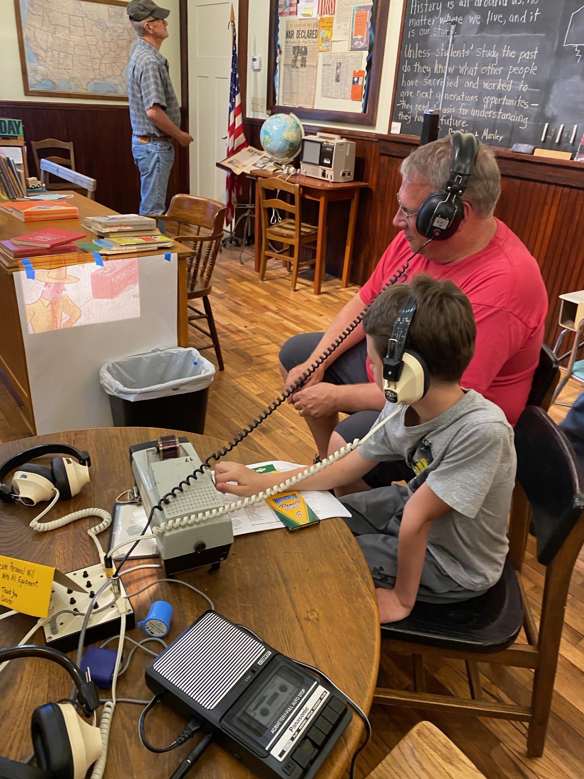 A boy and an adult wearing headphones operate a radio at a round table, another man stands nearby.
