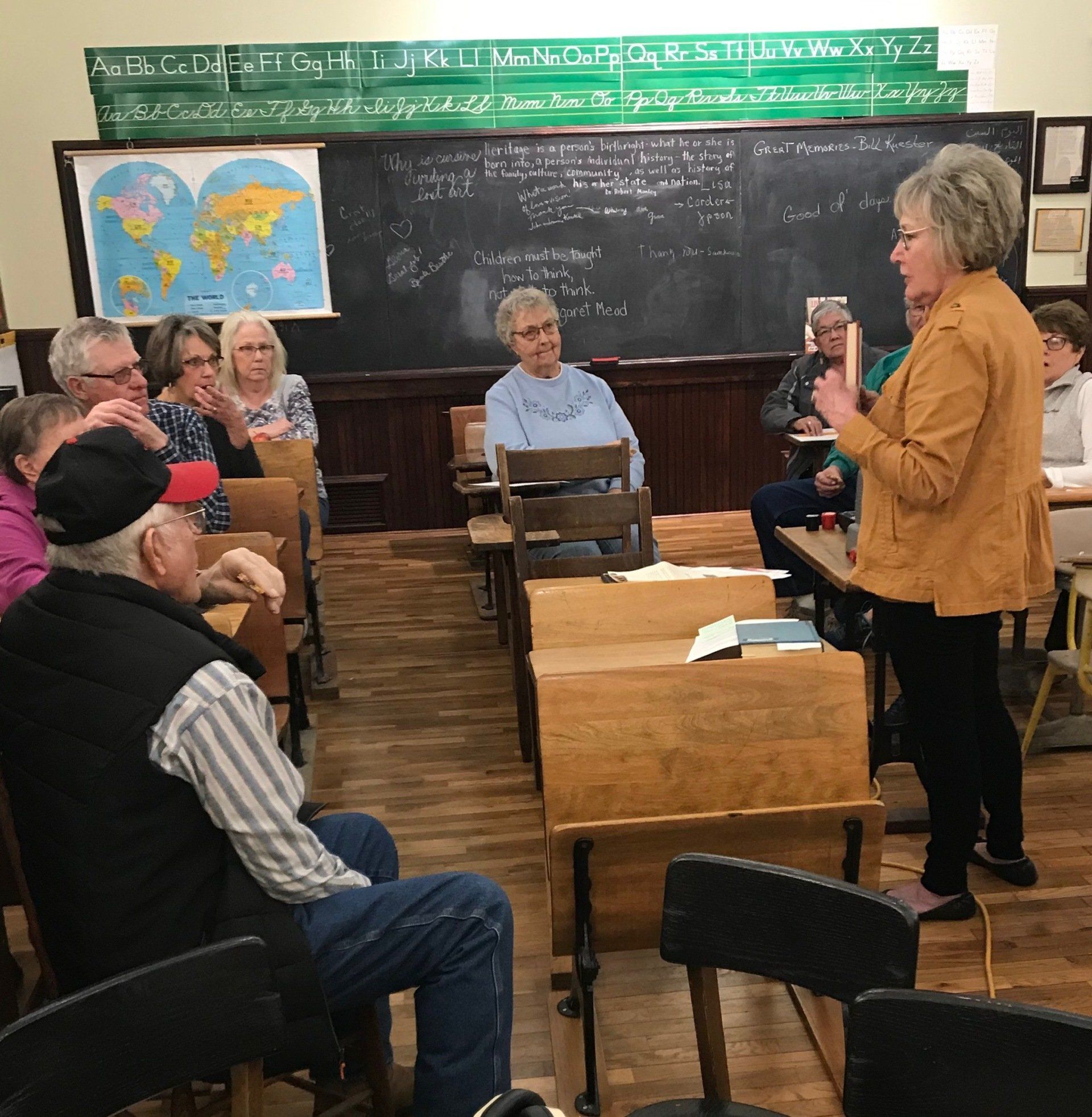 A woman speaks to a seated group in a vintage classroom.