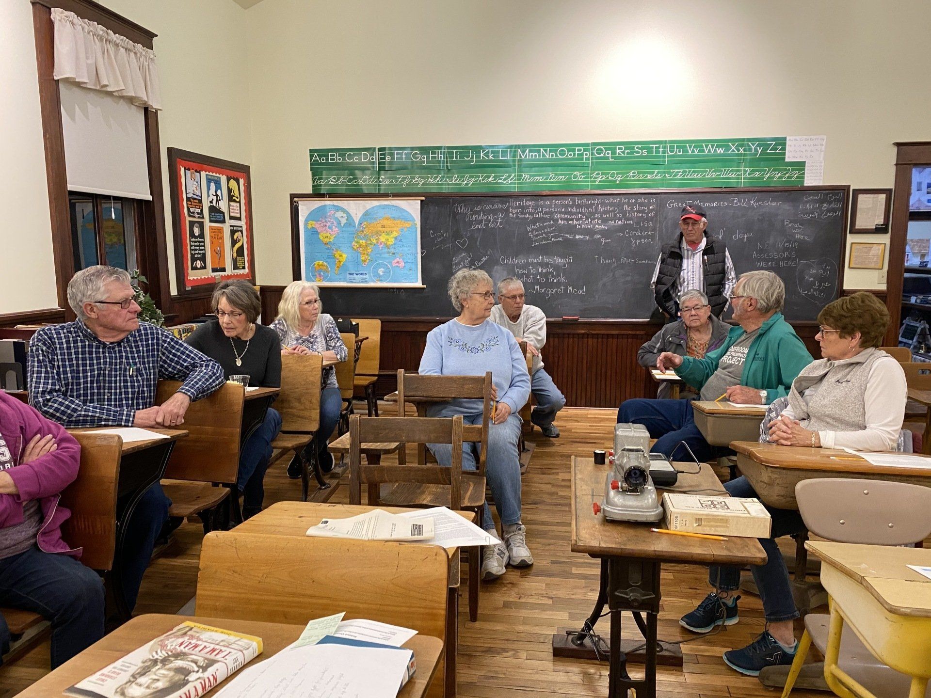 People sitting in a classroom, listening to a presentation. Wooden desks and chalkboard are visible.