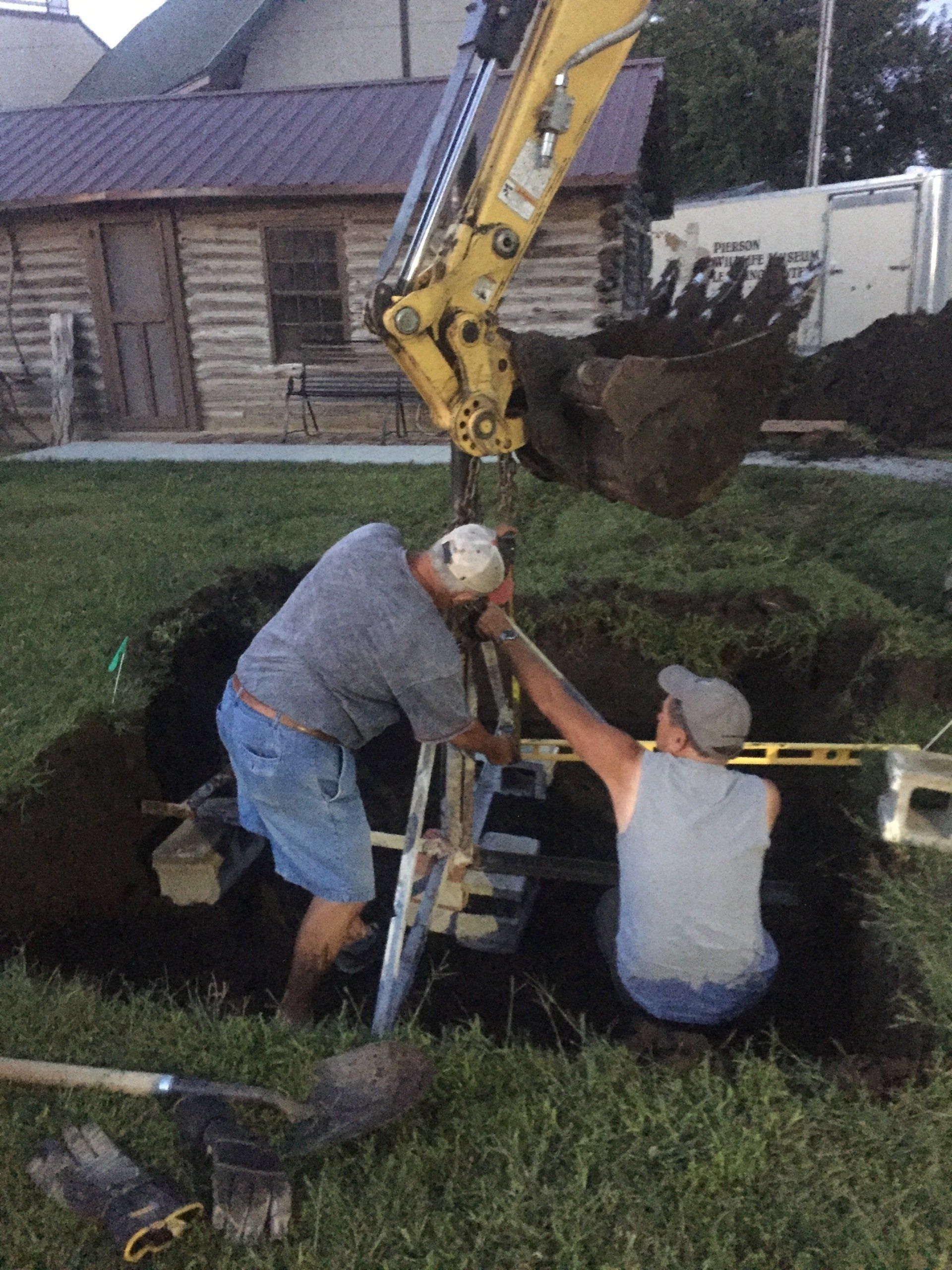 Two men working in a construction pit with an excavator arm above them. They use a level and ladder.