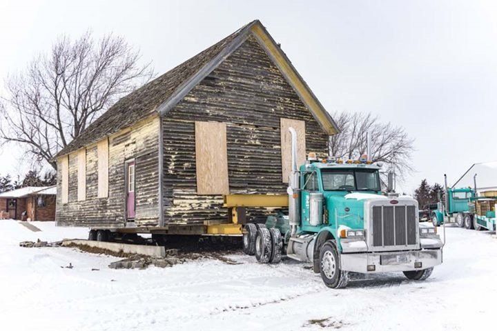 Old wooden church building being transported on a truck in a snowy landscape.
