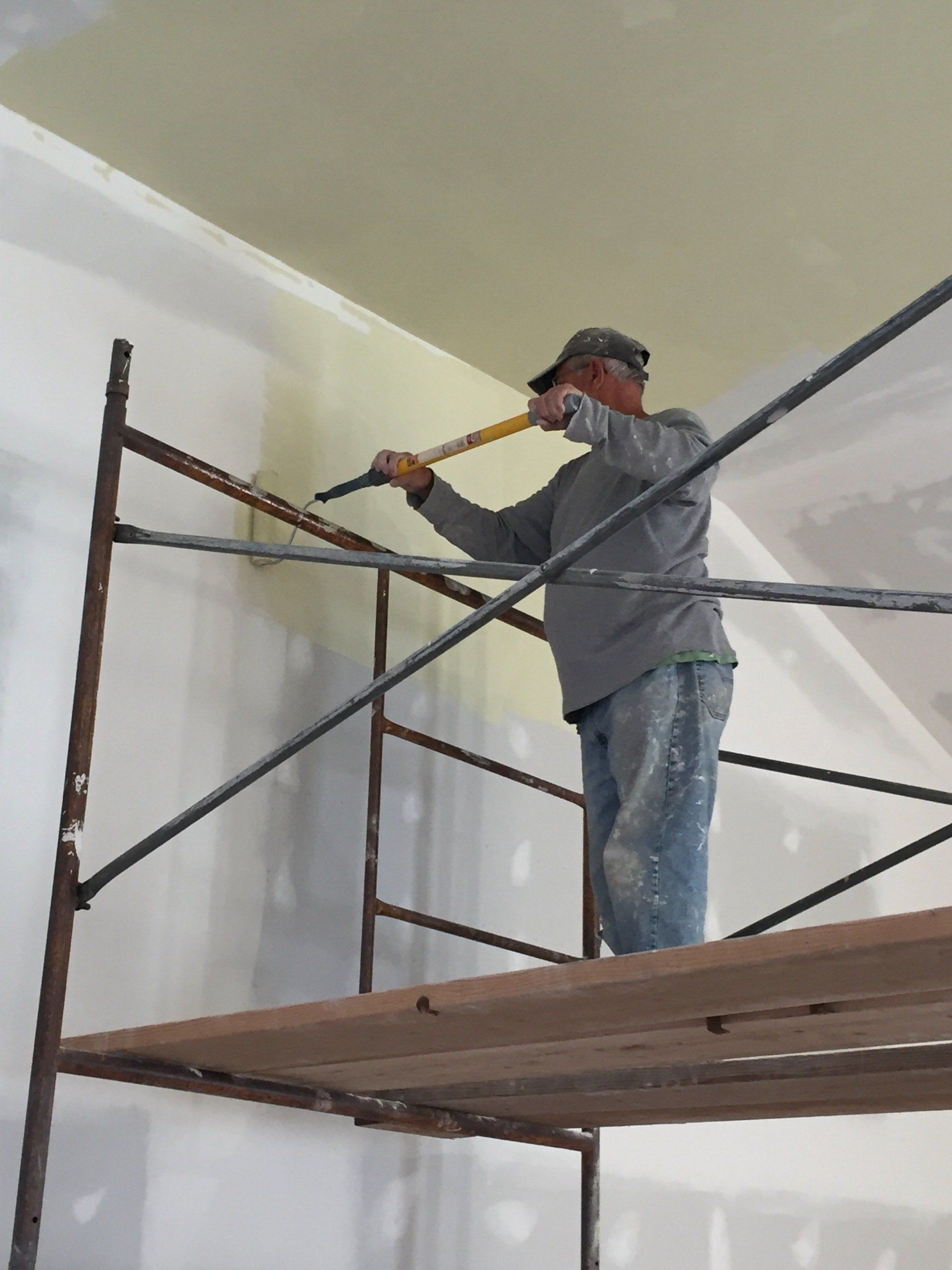 Person on scaffolding painting a ceiling, wearing a hat, gray shirt, and jeans. Interior setting.