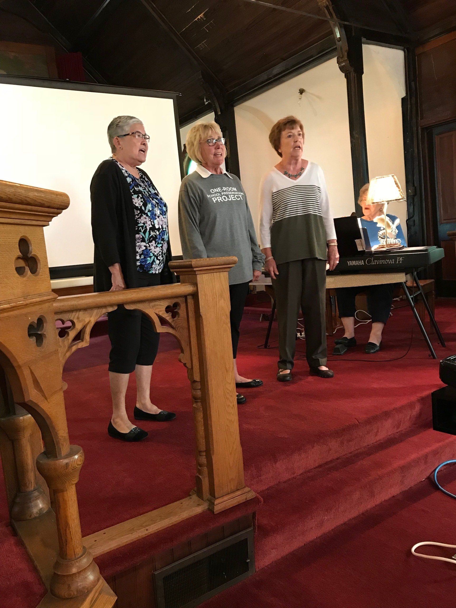 Three women singing on a stage with a keyboard player. Red carpet, wooden church interior.