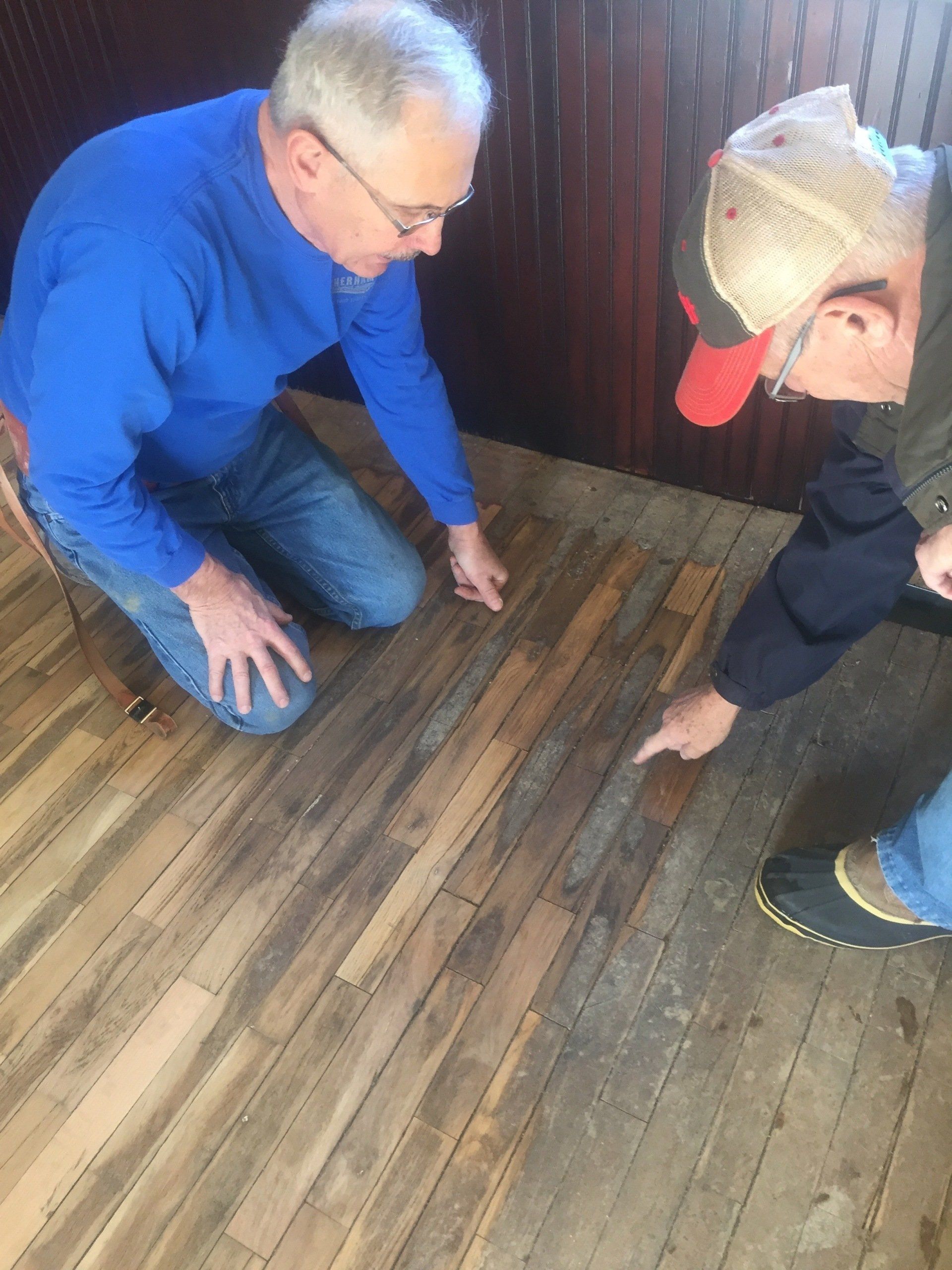 Two people examine a wooden floor, pointing. One kneels, wearing blue; the other wears a hat and jacket.
