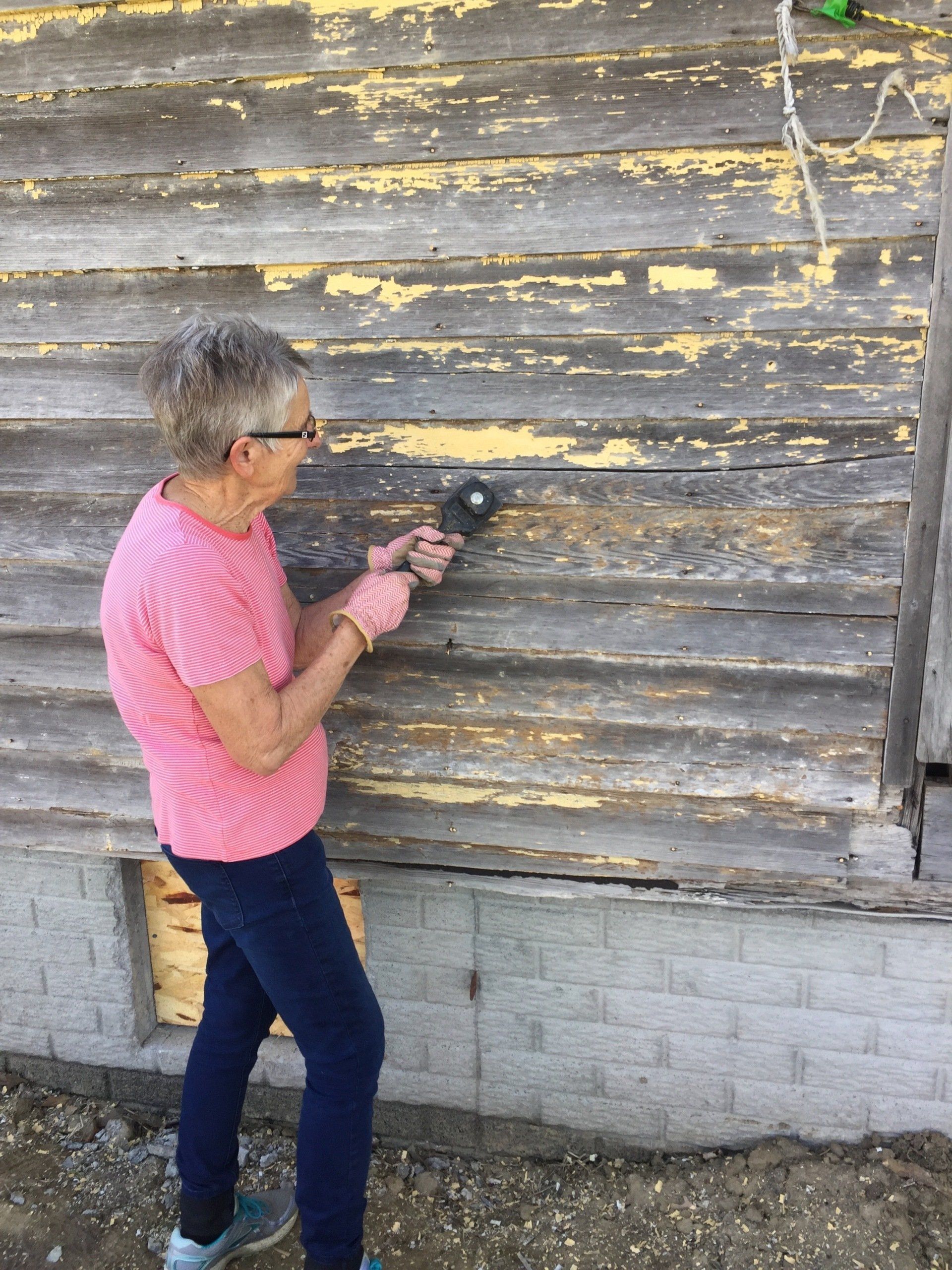 Woman using a tool to strip paint off of wooden siding on a building's exterior.