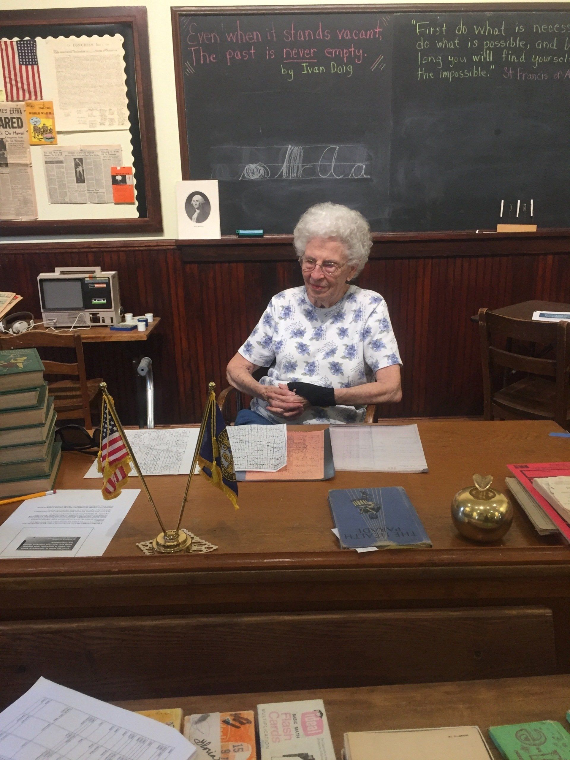 Woman seated at a wooden desk with papers, an old television, and a chalkboard in the background.