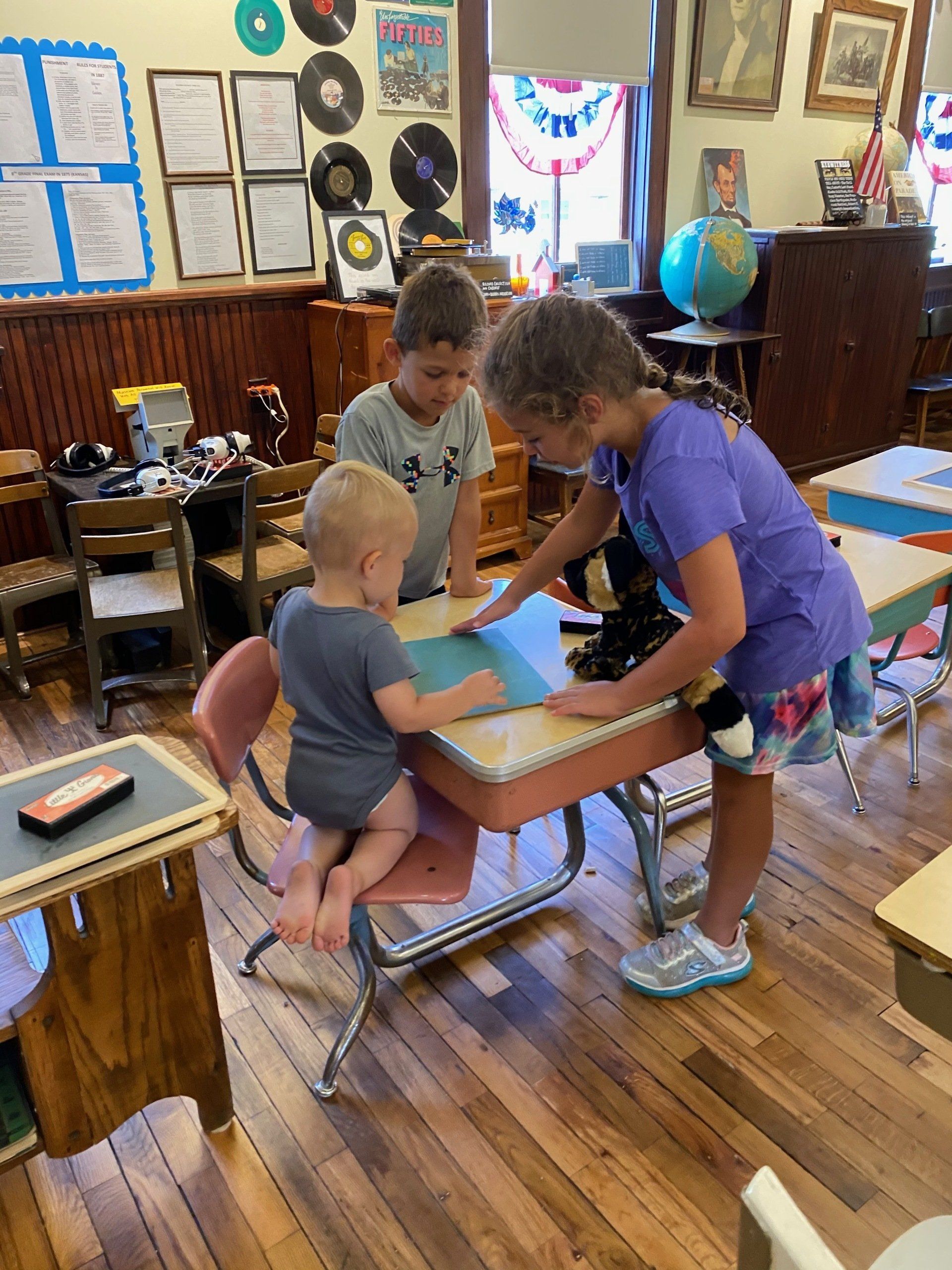 Children gather around a desk in a classroom, looking at a paper.