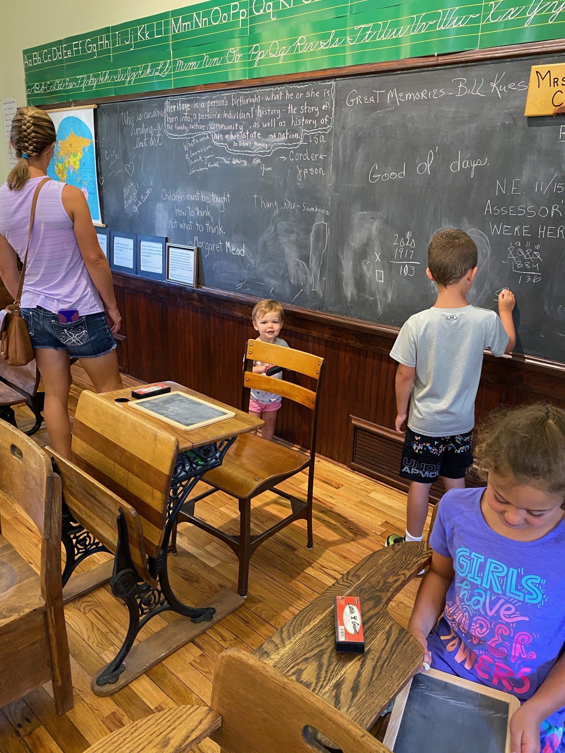 Children in a vintage classroom with a blackboard; one child writes math problems, another smiles.