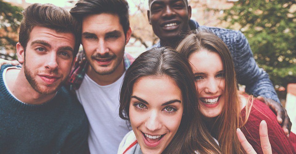 Group of friends smiling at the camera, outdoors. One woman takes a selfie.