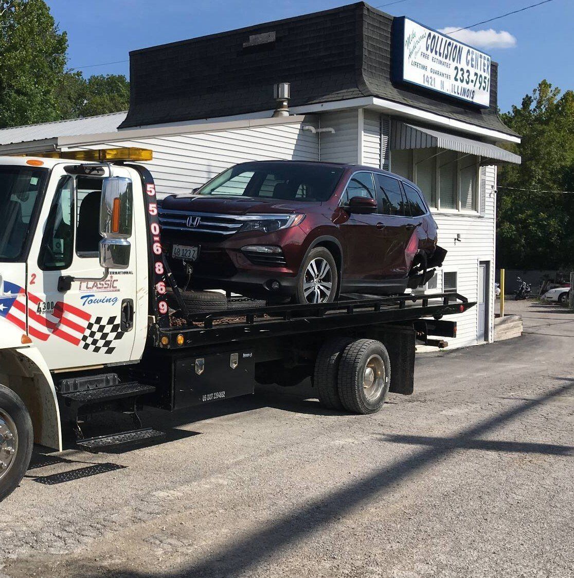 Tow truck hauling a maroon SUV beside a small white roadside building under a sunny sky