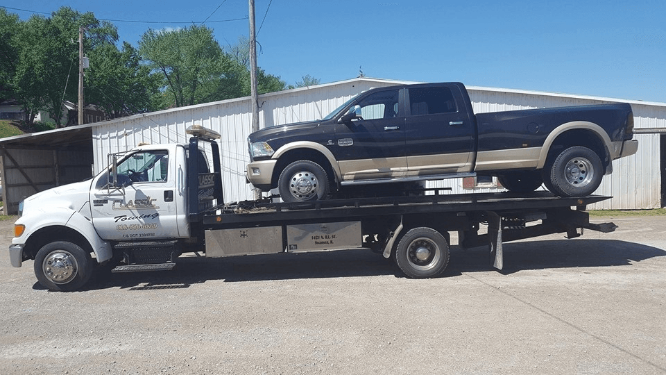 Tow truck carrying a black pickup on its flatbed, parked outdoors in bright sunlight