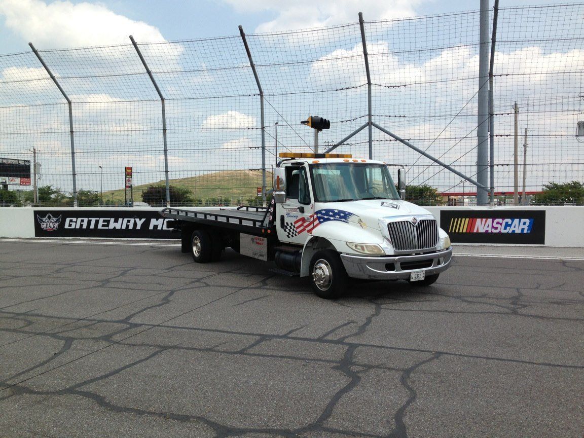 Tow truck on a racetrack beside safety fencing and sponsor banners under a cloudy sky